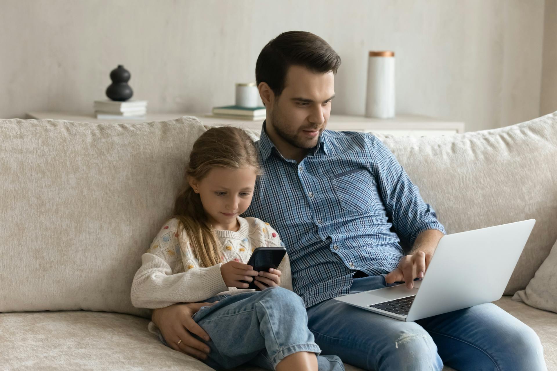 Mand sidder og kigger på sin computerskærm mens pige sidder ved siden af ham og kigger på sin telefon. De sidder tæt sammen i en sofa.