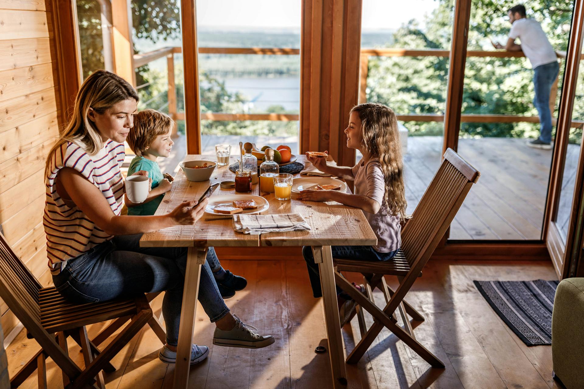 Kvinde kigger på sin telefon mens hun spiser morgenmad med sine børn. Børnene kigger smilende på hinanden.