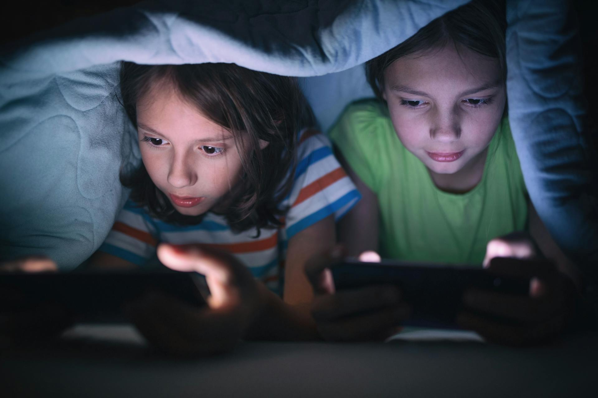 To piger ligger i mørket under tæppe og kigger på hver deres telefon.