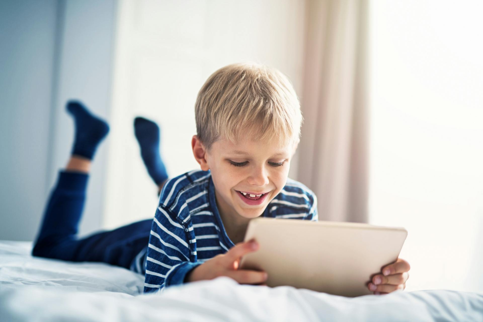 Dreng ligger i seng og kigger smilende ned på en tablet.