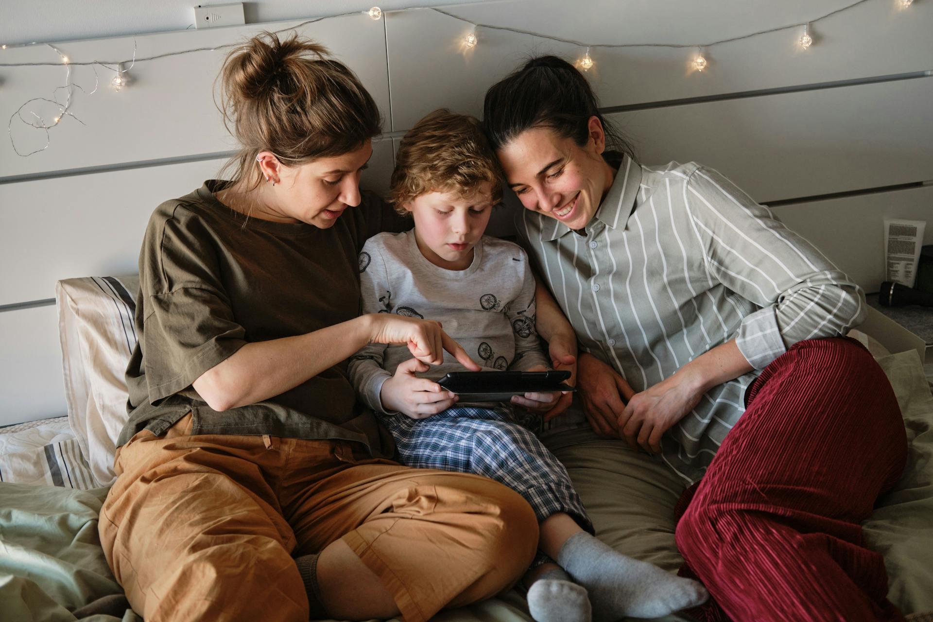 To kvinder ligger i en seng med en ung dreng imellem sig. De kigger alle tre på barnets tablet.
