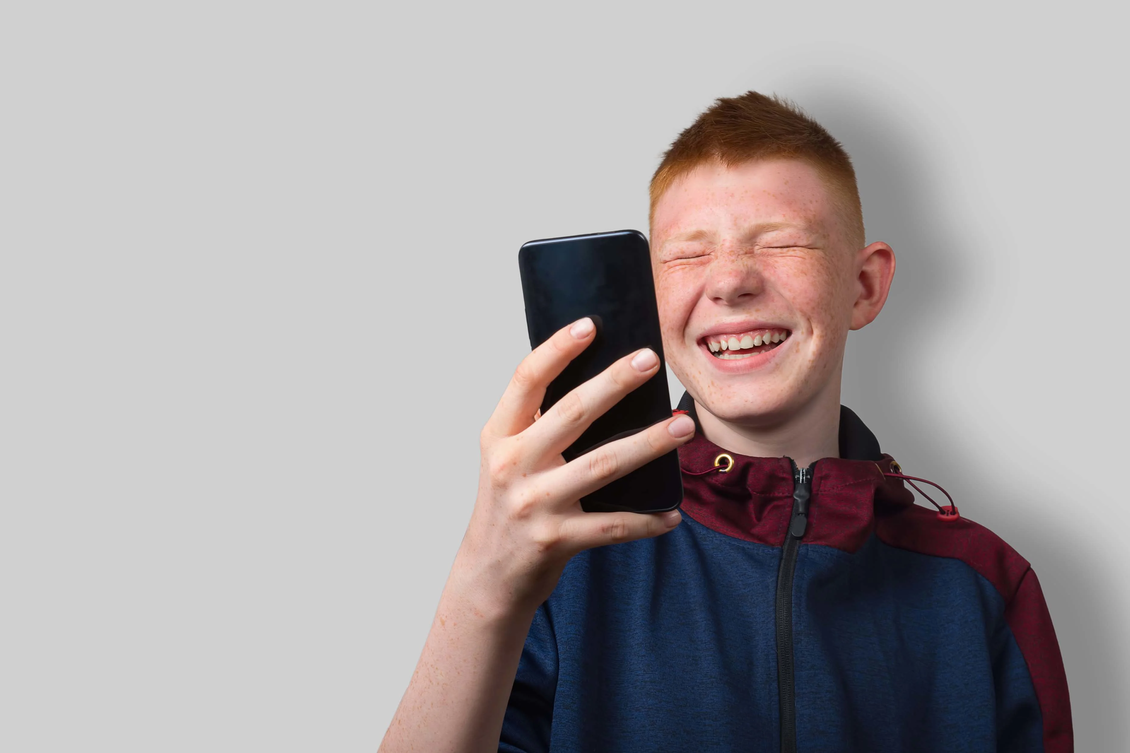Dreng griner med lukkede øjne med sin smartphone holdt op foran ansigtet.